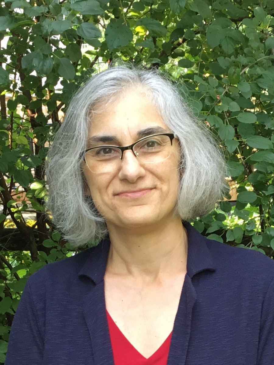 Christine Zaza stands in front of a leafy background. She wears glasses and has chin-length white hair. She wears a red shirt with a navy blazer on top. 